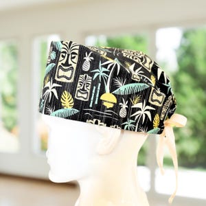 May include: A black surgical scrub cap with a tropical tiki and palm tree pattern. The cap features a repeating design of tiki masks, pineapples, palm trees, and a yellow sun. The cap has a light yellow tie at the back.