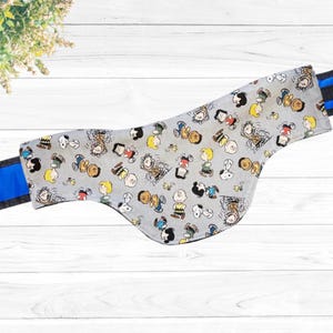 May include: A gray fabric dog bandana with a Peanuts character print. The bandana has a blue and black trim.