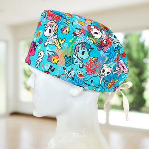 May include: A light blue surgical scrub cap featuring a colorful underwater scene with cartoon characters, including mermaids, sharks, and seahorses. The cap has a white tie closure and is designed for medical professionals or those in the healthcare field.