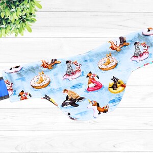 May include: A blue fabric dog bandana with a white background and a pattern of dogs and other animals on inner tubes and floaties. The bandana is folded in half and has a blue and white trim.