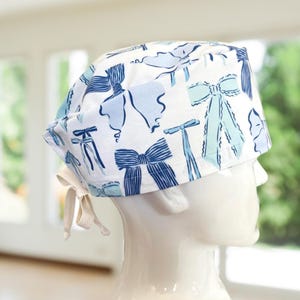 May include: A white surgical scrub cap with a repeating pattern of blue bows. The bows vary in style and shade, with some having striped details. The cap has a tie closure at the back, and is displayed on a white mannequin head.