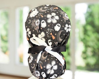 May include: A black and white patterned scrub hat featuring a whimsical design of ghosts, flowers, mushrooms, and crescent moons. The hat is tied with a black and white ribbon. The background is blurred, suggesting an indoor setting.