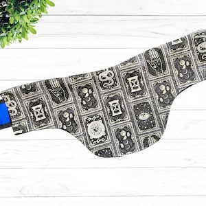 May include: A black fabric neck gaiter with a white and gold pattern of various symbols, including an eye, a snake, an hourglass, and a hand.