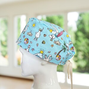 May include: A light blue surgical scrub cap featuring a whimsical beach-themed pattern. The design includes cartoon ducks, sandcastles, suns, seashells, and palm trees. The cap has a tie closure at the back and is displayed on a white mannequin head.