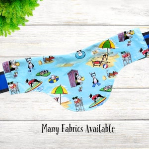 May include: A blue and white fabric dog bandana with a beach theme. The bandana features dogs wearing sunglasses, life jackets, and surfboards. The text "Many Fabrics Available" is printed at the bottom of the bandana.