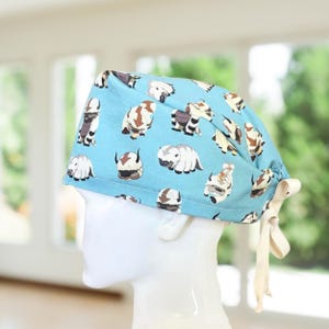 May include: A light blue scrub cap featuring a repeating pattern of white and brown cartoon bison characters with arrow markings on their heads. The cap includes a tie-back closure with cream-colored fabric ribbons at the rear for adjustment.