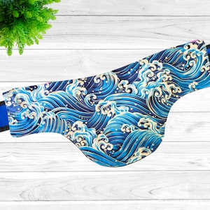 May include: A blue and gold patterned fabric item, possibly a pet accessory, featuring a repeating wave design. The item has a curved shape and a blue strap. The background is a white wooden surface.