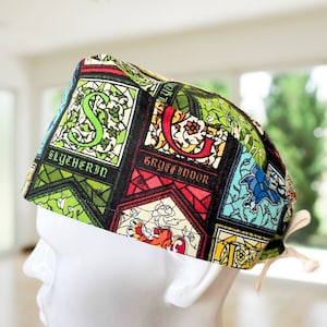 May include: A patterned surgical scrub cap featuring the words "Slytherin" and "Gryffindor" in a stained glass design. The cap is primarily green, red, and black, with a tie closure. The cap is on a white mannequin head.