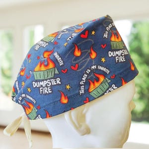May include: A blue surgical scrub cap with a repeating pattern of burning dumpsters, hearts, stars, and the text "DUMPSTER FIRE" and "THIS PLACE IS MY FAVORITE". The cap has a cream-colored tie.