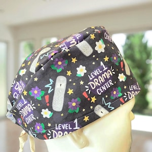 May include: Black scrub hat with a white background and a repeating pattern of white stars, purple flowers, and white outlines of a medical device. The text "Level 1 Drama Center" is repeated throughout the pattern.