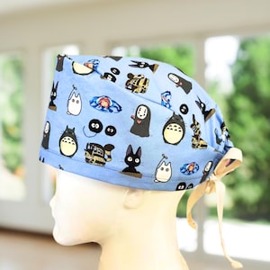 May include: A light blue scrub cap featuring a repeating pattern of various animated characters including a black cat, a white creature, a dark spirit, and soot sprites. The cap has a tie-back closure with beige fabric ribbons at the rear.