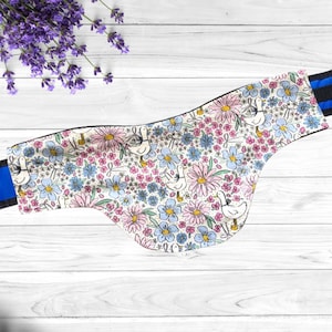 May include: A floral patterned horse saddle pad featuring a white background with pink and blue flowers and white ducks. The pad has a black and blue striped trim and is set against a white wooden background.
