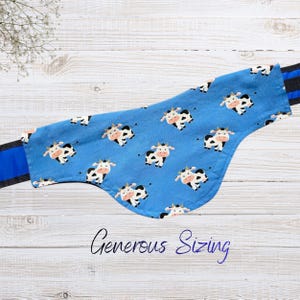 May include: A blue pet bandana with a repeating pattern of black and white cartoon cows. The bandana has a curved shape and is trimmed with a black and blue striped fabric. The text "Generous Sizing" is written in cursive at the bottom.