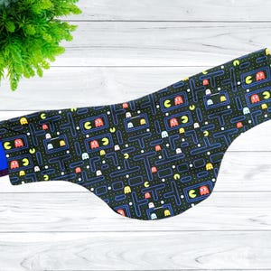 May include: A black fabric dog bandana with a Pac-Man pattern. The bandana features a maze with Pac-Man ghosts and pellets in various colors, including red, yellow, blue, and pink.