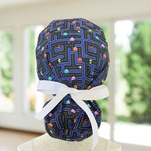 May include: A blue and black scrub hat with a white tie. The hat features a Pac-Man maze pattern with colorful Pac-Man characters.