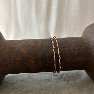 K&ouml;nnte beinhalten: Zwei zarte Armb&auml;nder mit Kupfer- und wei&szlig;en Perlen. Die Armb&auml;nder sind auf einer rostigen Metalloberfl&auml;che &uuml;bereinander gelegt.
