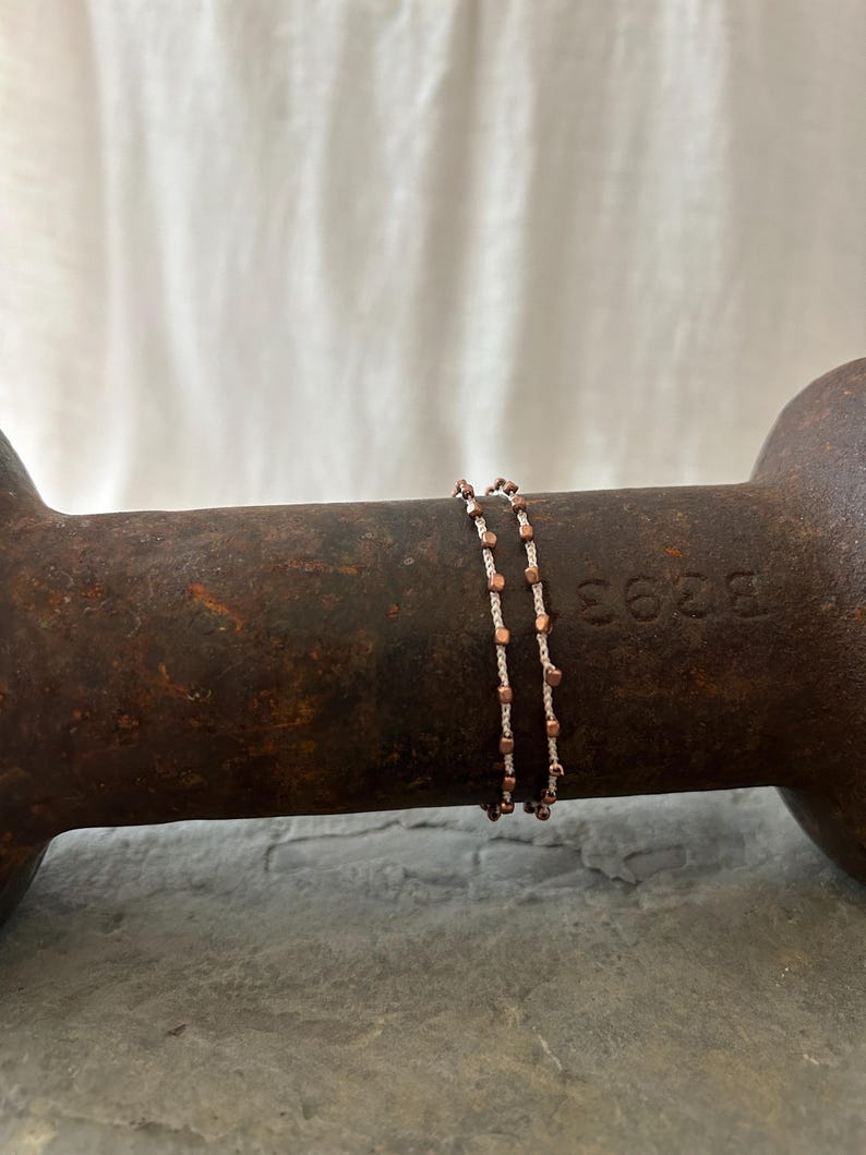 K&ouml;nnte beinhalten: Zwei zarte Armb&auml;nder mit Kupfer- und wei&szlig;en Perlen. Die Armb&auml;nder sind auf einer rostigen Metalloberfl&auml;che &uuml;bereinander gelegt.