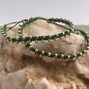 May include: Two green friendship bracelets with silver beads. The bracelets are tied together and are on a white rock.