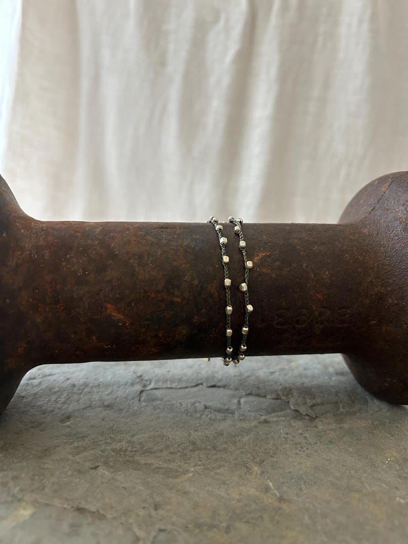 K&ouml;nnte beinhalten: Zwei silberne Kettenarmb&auml;nder mit kleinen quadratischen Perlen. Die Armb&auml;nder liegen &uuml;bereinander und ruhen auf einer braunen Oberfl&auml;che.