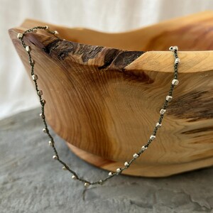 K&ouml;nnte beinhalten: Eine silberne und gr&uuml;ne Perlenkette mit einer zarten Kette. Die Kette liegt auf einer Holzschale.