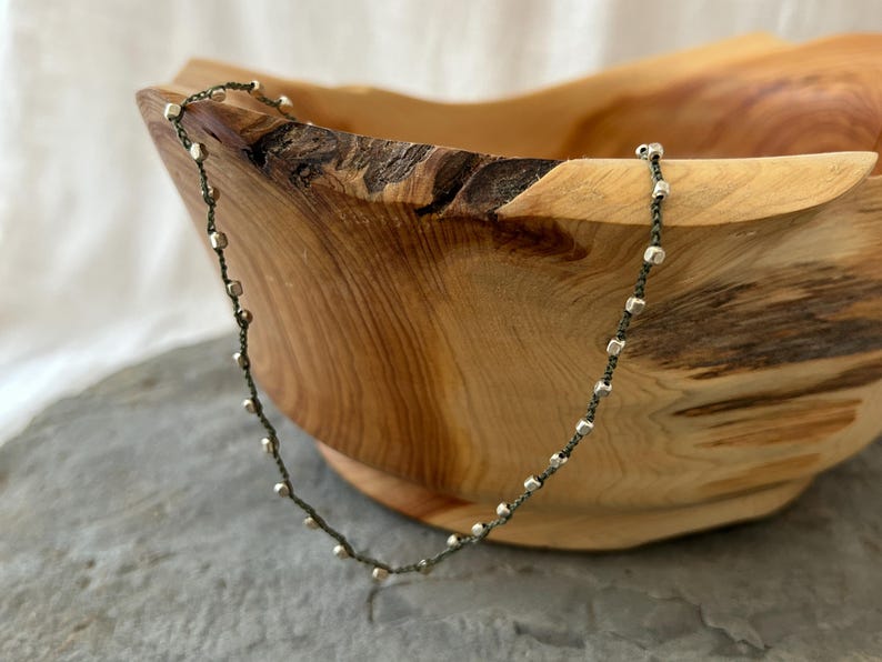 K&ouml;nnte beinhalten: Eine silberne und gr&uuml;ne Perlenkette mit einer zarten Kette. Die Kette liegt auf einer Holzschale.