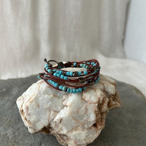 May include: A multi-strand wrap bracelet with turquoise beads and brown leather cords. The bracelet features small, square metal accents and a clasp closure. The jewelry is displayed on a white and gray stone.