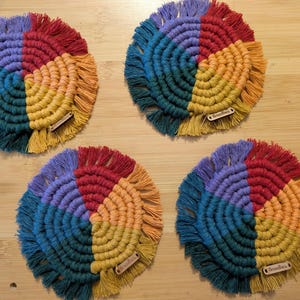 Könnte beinhalten: Vier handgefertigte Makramee-Untersetzer in Regenbogenfarben. Jeder Untersetzer ist in vier Farbbereiche unterteilt: Blau, Lila, Rot und Gelb, mit einem Fransenrand. An jedem Untersetzer sind kleine Holzanhänger befestigt.