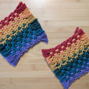 Könnte beinhalten: Zwei quadratische Makramee-Untersetzer in Regenbogenfarben. Die Untersetzer haben ein gewebtes Design mit Fransen an den Kanten. Die Farben wechseln von Rot über Orange, Gelb, Grün, Blau bis hin zu Violett.