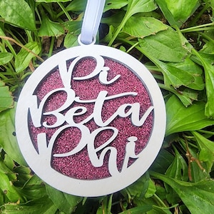 May include: A circular, silver-toned ornament featuring the cut-out text "Pi Beta Phi". The centre is filled with a sparkly, magenta material. A white ribbon is attached for hanging. The ornament is displayed on a bed of green leaves.