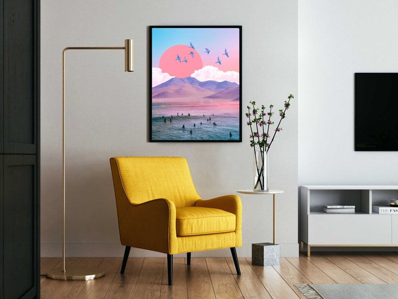 May include: A framed print of a surreal landscape with a pink sun, mountains, and a beach with people in the water. The print is in a black frame and is hanging on a white wall.