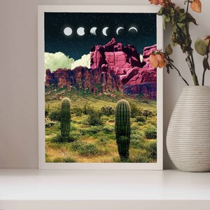 May include: A framed print of a desert landscape with two tall cacti in the foreground. The background features a mountain range with a pink hue and a night sky with a crescent moon and stars.