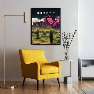 May include: A framed print of a desert landscape with a pink mountain range, cacti, and a crescent moon in the sky. The print is hanging on a white wall in a living room setting.