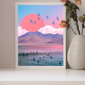 May include: A framed print of a surreal landscape with a pink sun, blue birds, and a group of surfers in the water. The mountains in the background are purple and the sky is blue.