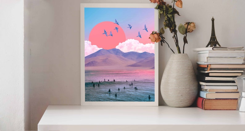 May include: A framed print of a surreal landscape with a pink sun, blue birds, and a group of surfers in the water. The mountains in the background are purple and the sky is blue.