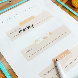 May include: A clear plastic sheet with a teal border, featuring a planner layout. The text "Days of the Week" is visible. The word "Monday" is written on a beige label. Decorative floral tape accents the labels.