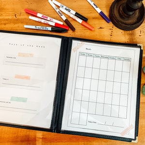 May include: A black binder with two pages, one labeled "Days of the Week" and the other "Month". The "Days of the Week" page has three blank lines with spaces for writing. The "Month" page has a calendar grid with spaces for writing the days of the week and dates. The binder is open and lying on a wooden surface.