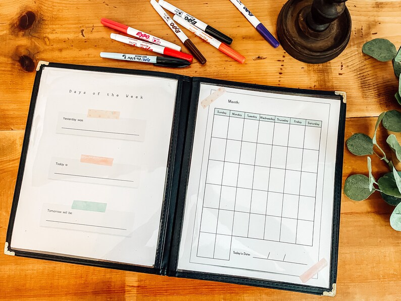 May include: A black binder with two pages, one labeled "Days of the Week" and the other "Month". The "Days of the Week" page has three blank lines with spaces for writing. The "Month" page has a calendar grid with spaces for writing the days of the week and dates. The binder is open and lying on a wooden surface.