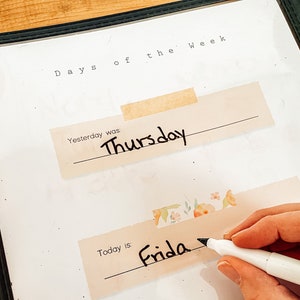 May include: A clear plastic dry erase board with the words "Days of the Week" at the top. The word "Thursday" is written on a beige label, and the word "Friday" is being written with a black marker. The board is in a black frame.