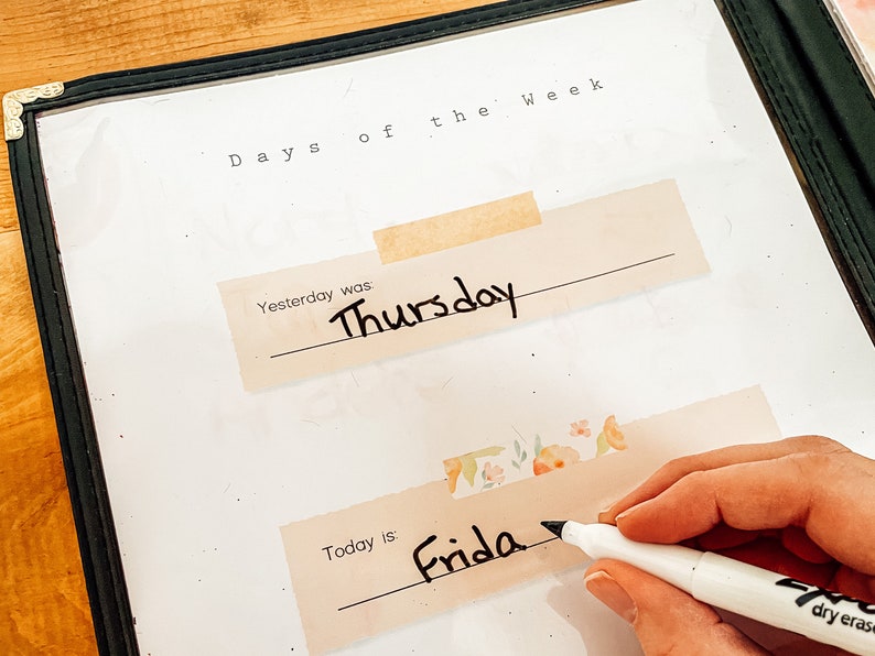 May include: A clear plastic dry erase board with the words "Days of the Week" at the top. The word "Thursday" is written on a beige label, and the word "Friday" is being written with a black marker. The board is in a black frame.