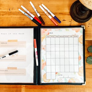 May include: A black binder with a clear plastic cover, containing a calendar and a "Days of the Week" section. The calendar has a floral design. Several colored markers are scattered around the binder on a wooden surface.
