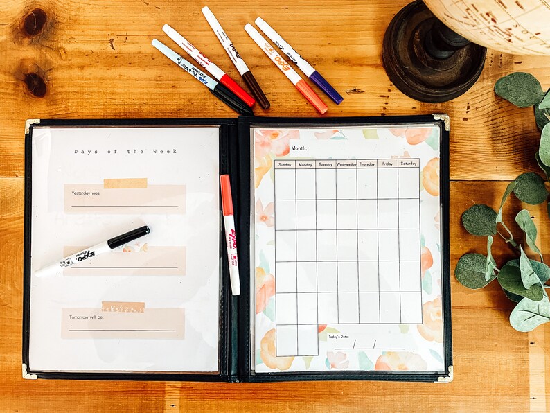 May include: A black binder with a clear plastic cover, containing a calendar and a "Days of the Week" section. The calendar has a floral design. Several colored markers are scattered around the binder on a wooden surface.