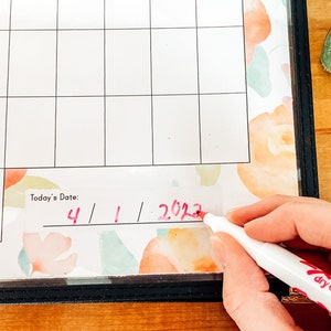 May include: A close-up of a white dry-erase calendar with a floral design. The date "4/1/2023" is being written in red marker. The calendar is in a black frame.