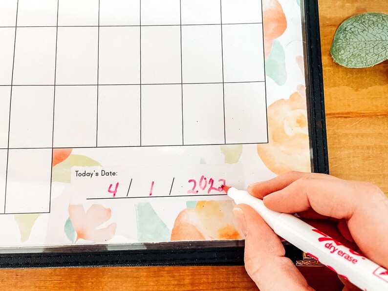 May include: A close-up of a white dry-erase calendar with a floral design. The date "4/1/2023" is being written in red marker. The calendar is in a black frame.