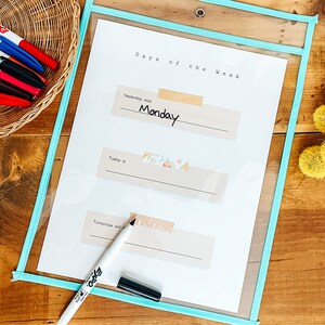 May include: A clear plastic pocket with a light blue border holds a white sheet labeled "Days of the Week." The word "Monday" is written on the sheet. A black marker and a basket of colorful markers are also visible.