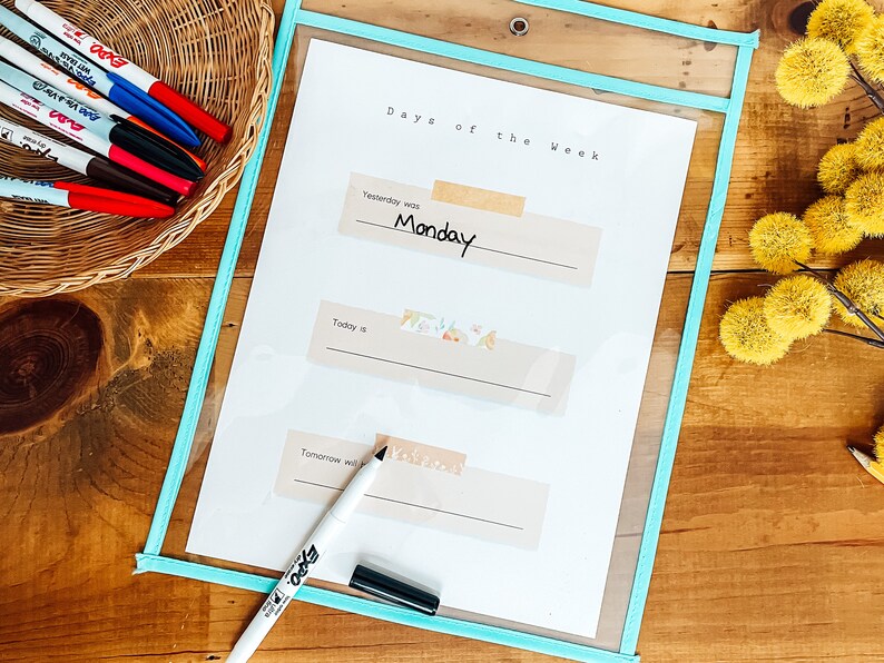 May include: A clear plastic pocket with a light blue border holds a white sheet labeled "Days of the Week." The word "Monday" is written on the sheet. A black marker and a basket of colorful markers are also visible.