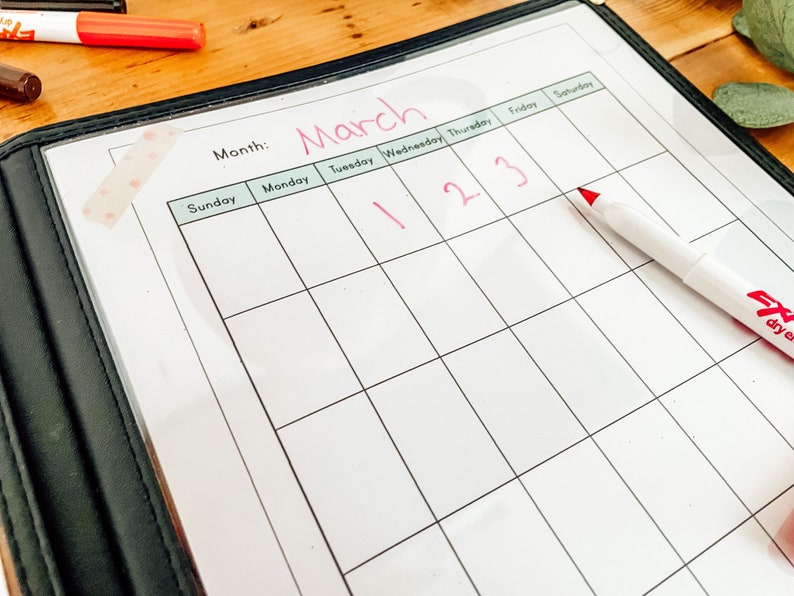 May include: A white dry-erase calendar with the month of March written at the top. The days of the week are listed across the top, and the numbers 1, 2, and 3 are written in red on the calendar.