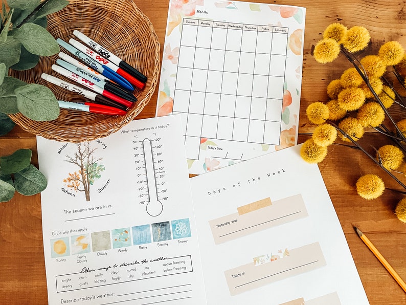 May include: A collection of educational printables, including a calendar, weather chart, and a 'Days of the Week' worksheet. A wicker basket holds colorful markers. Yellow pom-pom flowers and a pencil are also present.