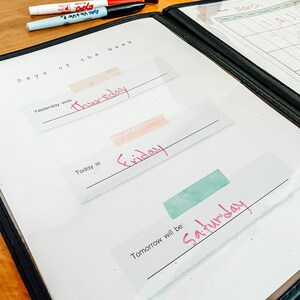 May include: A white dry-erase board with a black binder. The board has the text "Days of the Week" written at the top. There are three blank lines with the words "Yesterday was", "Today is", and "Tomorrow will be:" written above each line. The words "Thursday", "Friday", and "Saturday" are written in pink on the lines.