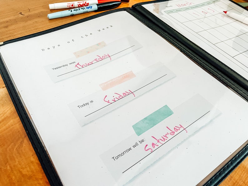 May include: A white dry-erase board with a black binder. The board has the text "Days of the Week" written at the top. There are three blank lines with the words "Yesterday was", "Today is", and "Tomorrow will be:" written above each line. The words "Thursday", "Friday", and "Saturday" are written in pink on the lines.