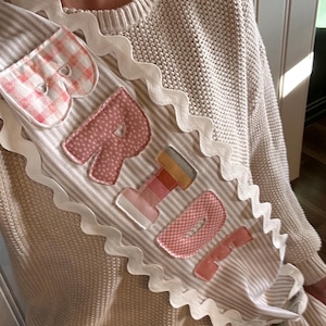 May include: A striped sash with the word "Bride" appliqued in pink fabric letters. The sash has a scalloped white trim and is designed to be worn over the shoulder. The letters are in various patterns.